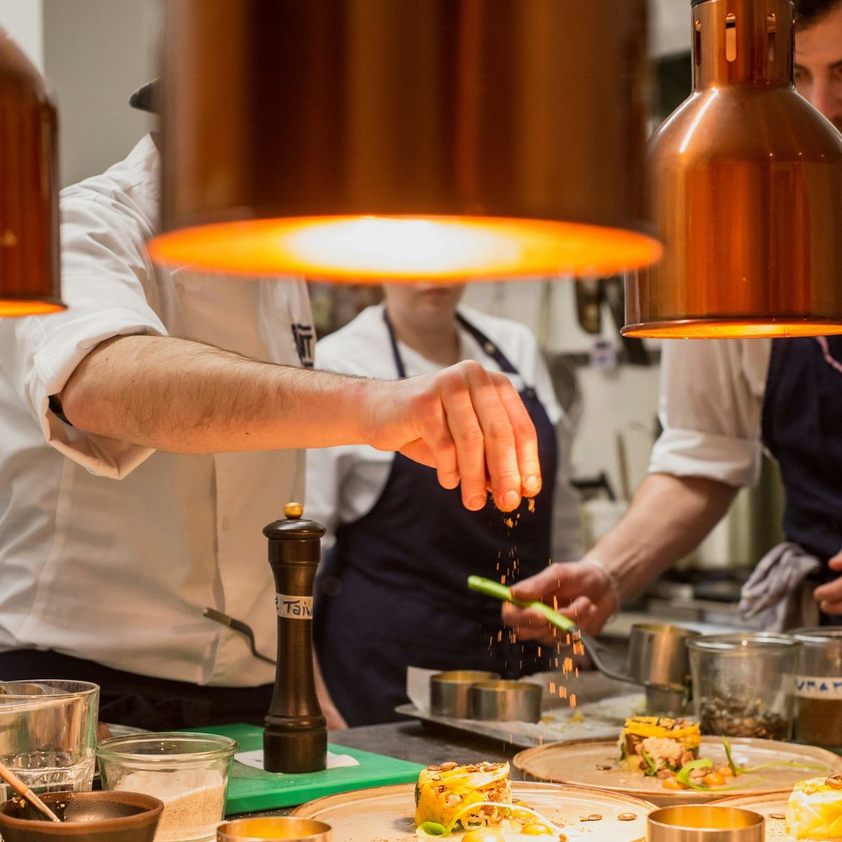 Chefs finishing plates beneath copper heat lamps in a professional kitchen