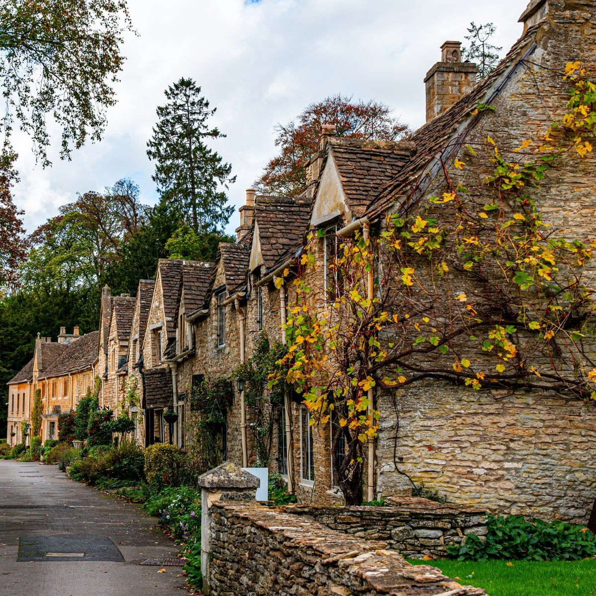 A row of honey-coloured stone cottages along a quiet Cotswolds lane, autumn creeper on the walls