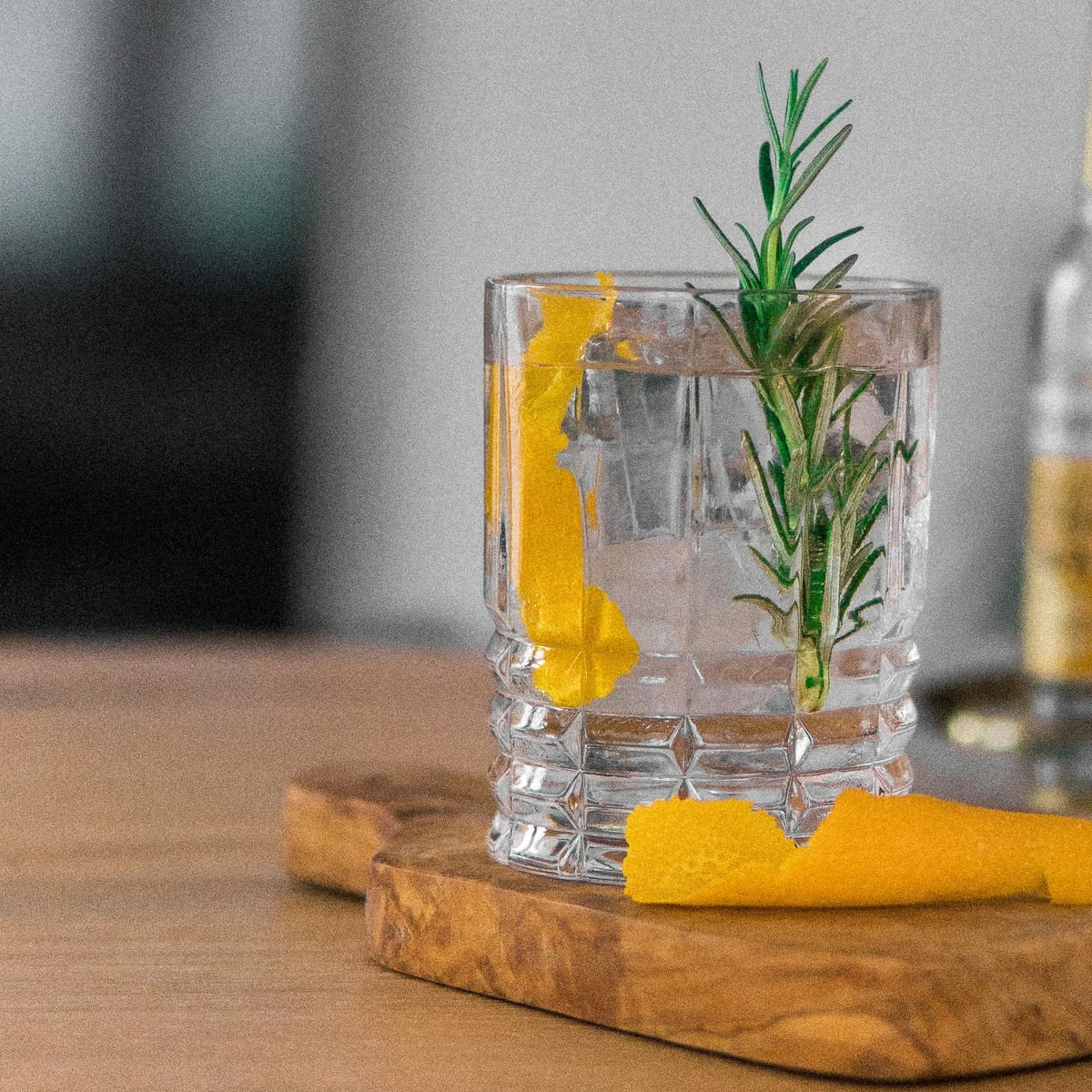 A gin and tonic with rosemary and orange peel in a cut-glass tumbler on an olive-wood board