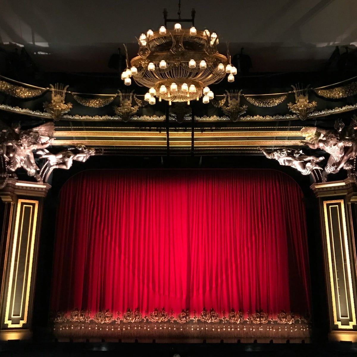 A theatre stage with its red curtain drawn, framed in gilt beneath a chandelier