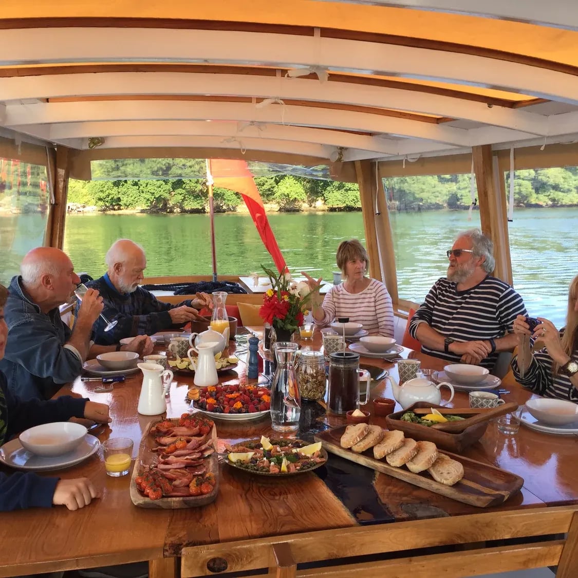 Guests at a long lunch table aboard a covered wooden boat, a wooded riverbank beyond the windows