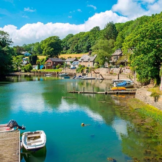 Boats moored on a still, tree-lined creek, stone cottages along the water's edge