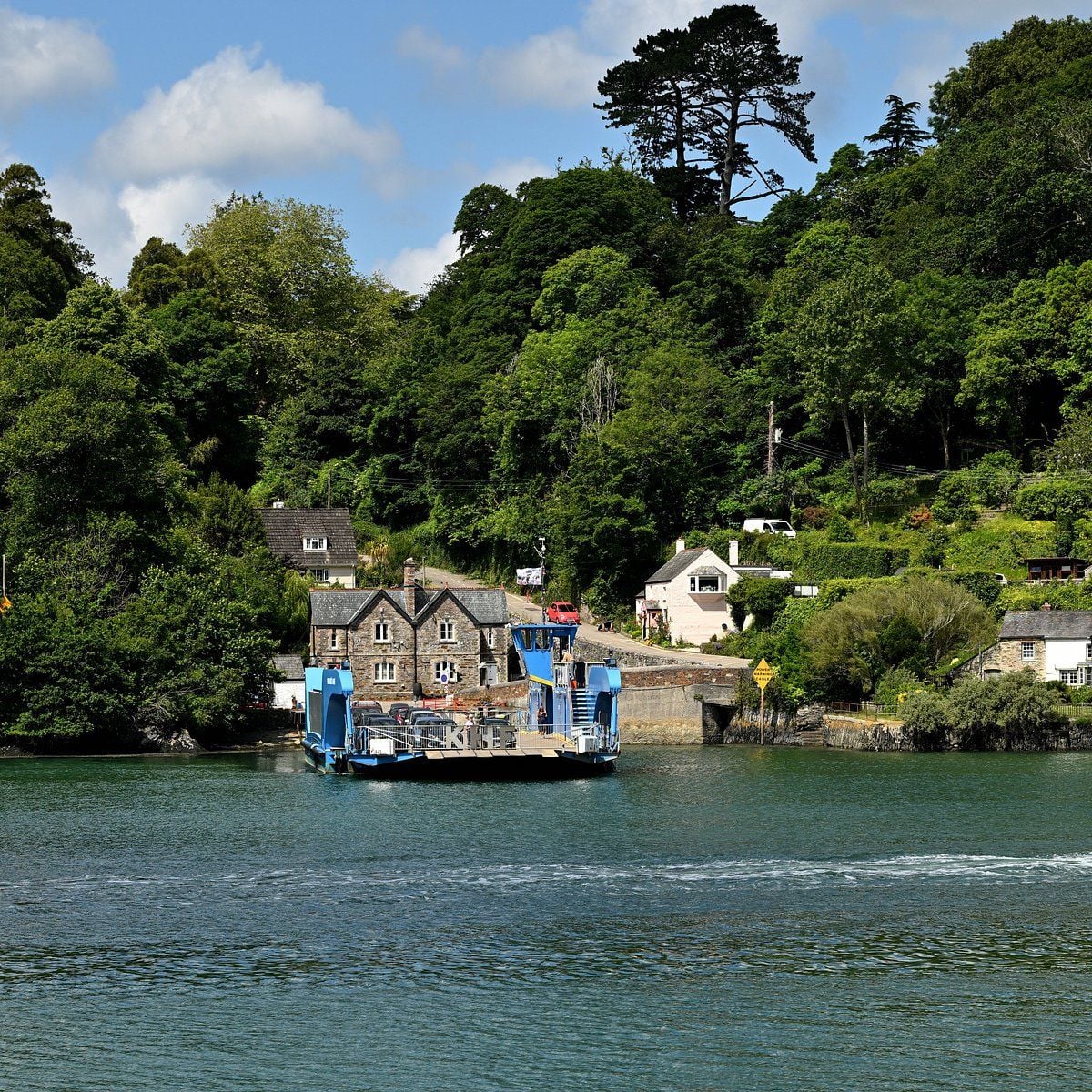 The blue King Harry chain ferry at its slipway on the River Fal, wooded banks and stone cottages behind