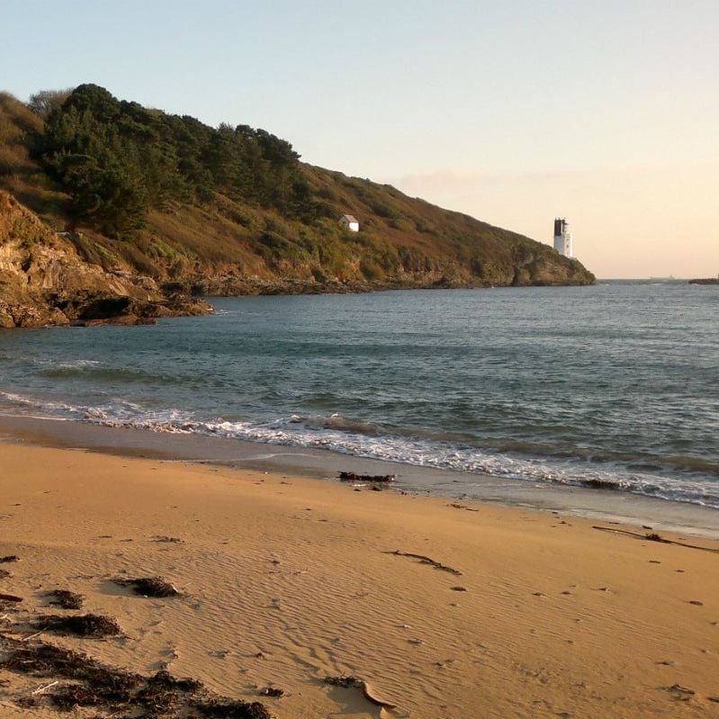 A sandy cove beneath low cliffs at evening, the white lighthouse on St Anthony Head in the distance