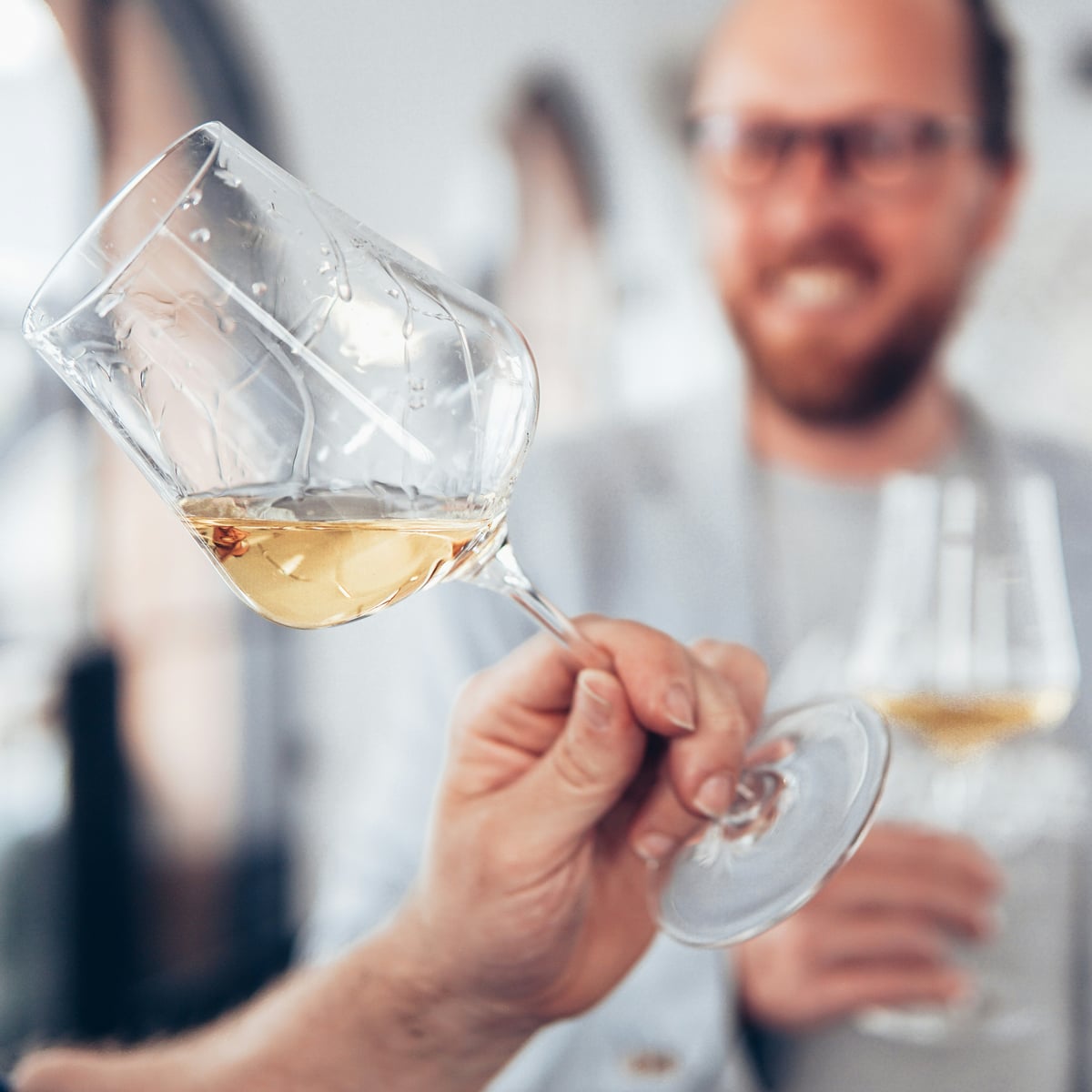 A glass of white wine held up to the light at a tasting, a smiling guest with his own glass behind