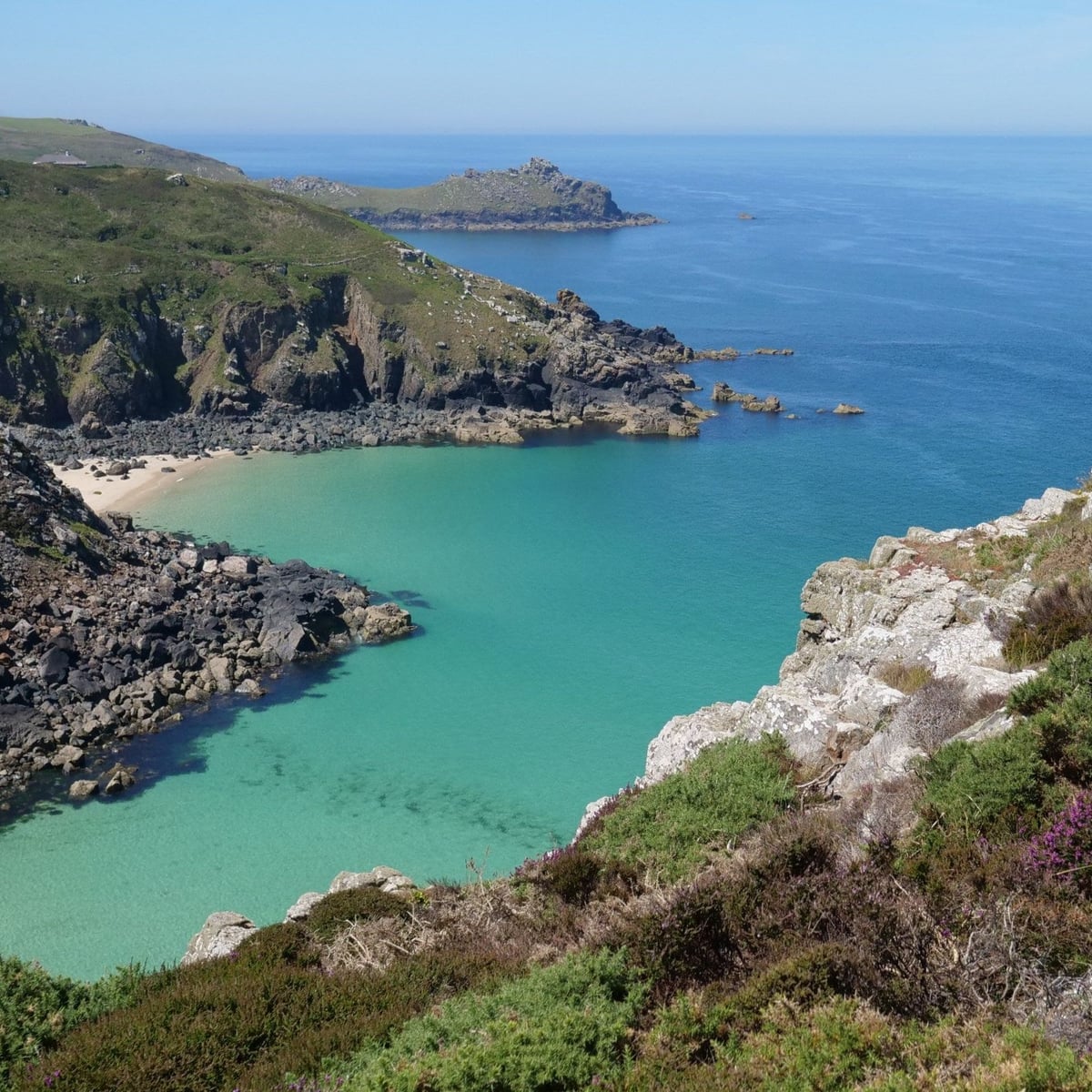 A turquoise cove below granite outcrops and heather on the coast path, a headland reaching out to sea beyond