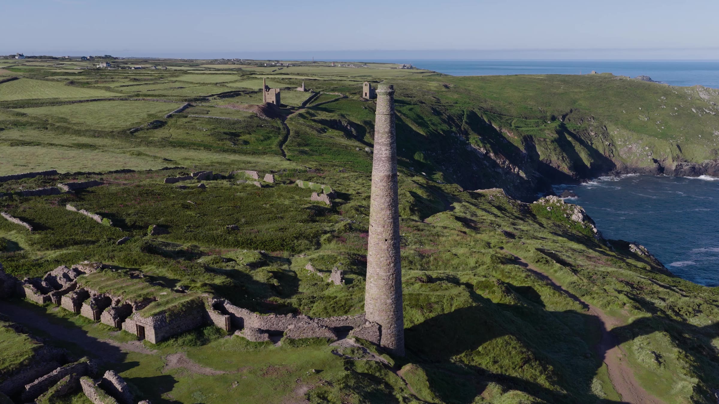 A mine chimney stack and ruined engine house walls on a green clifftop plateau above the sea at Botallack